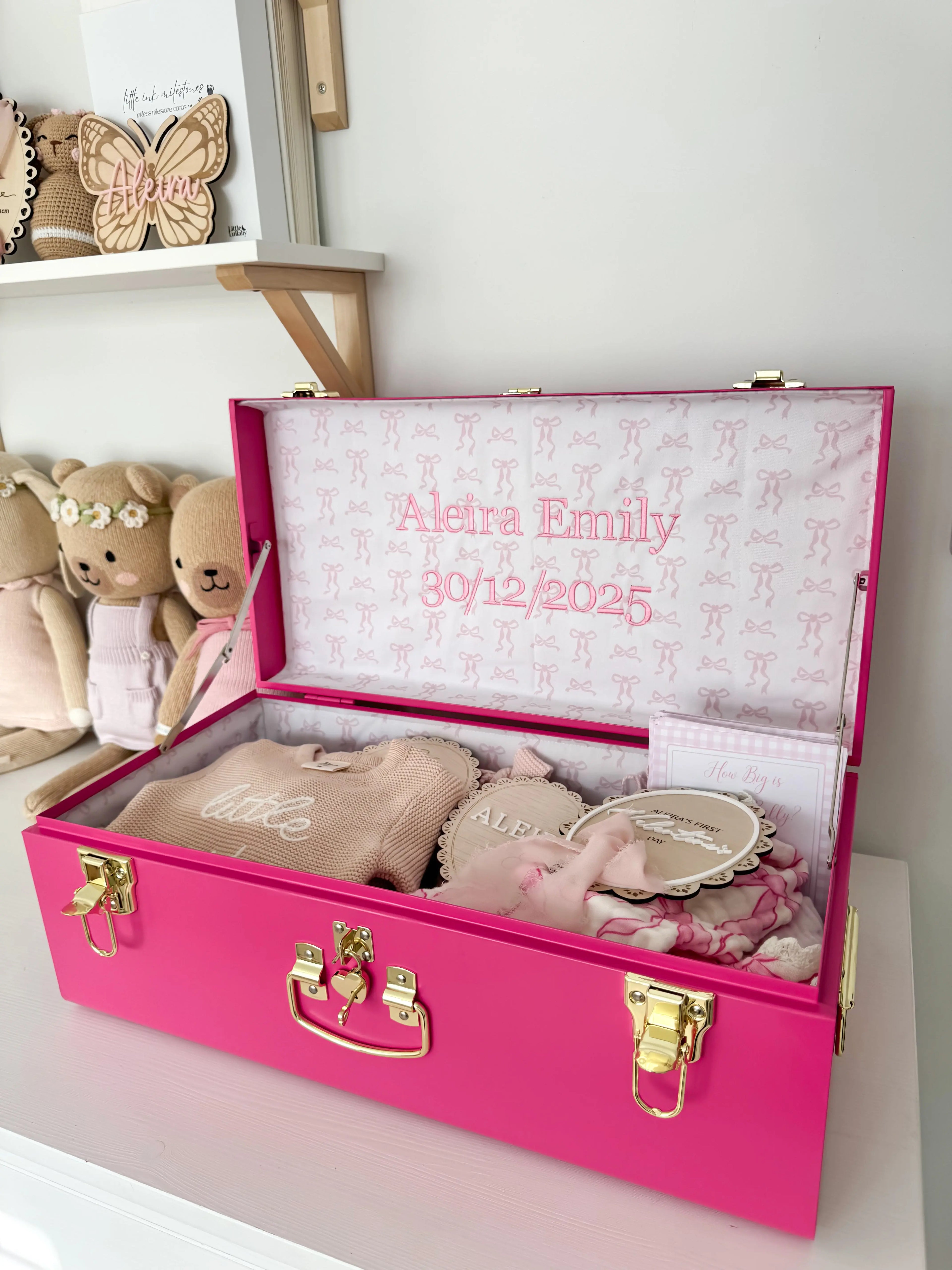 Pink baby trunk with embroidered name and date, surrounded by teddy bears and decorative items.