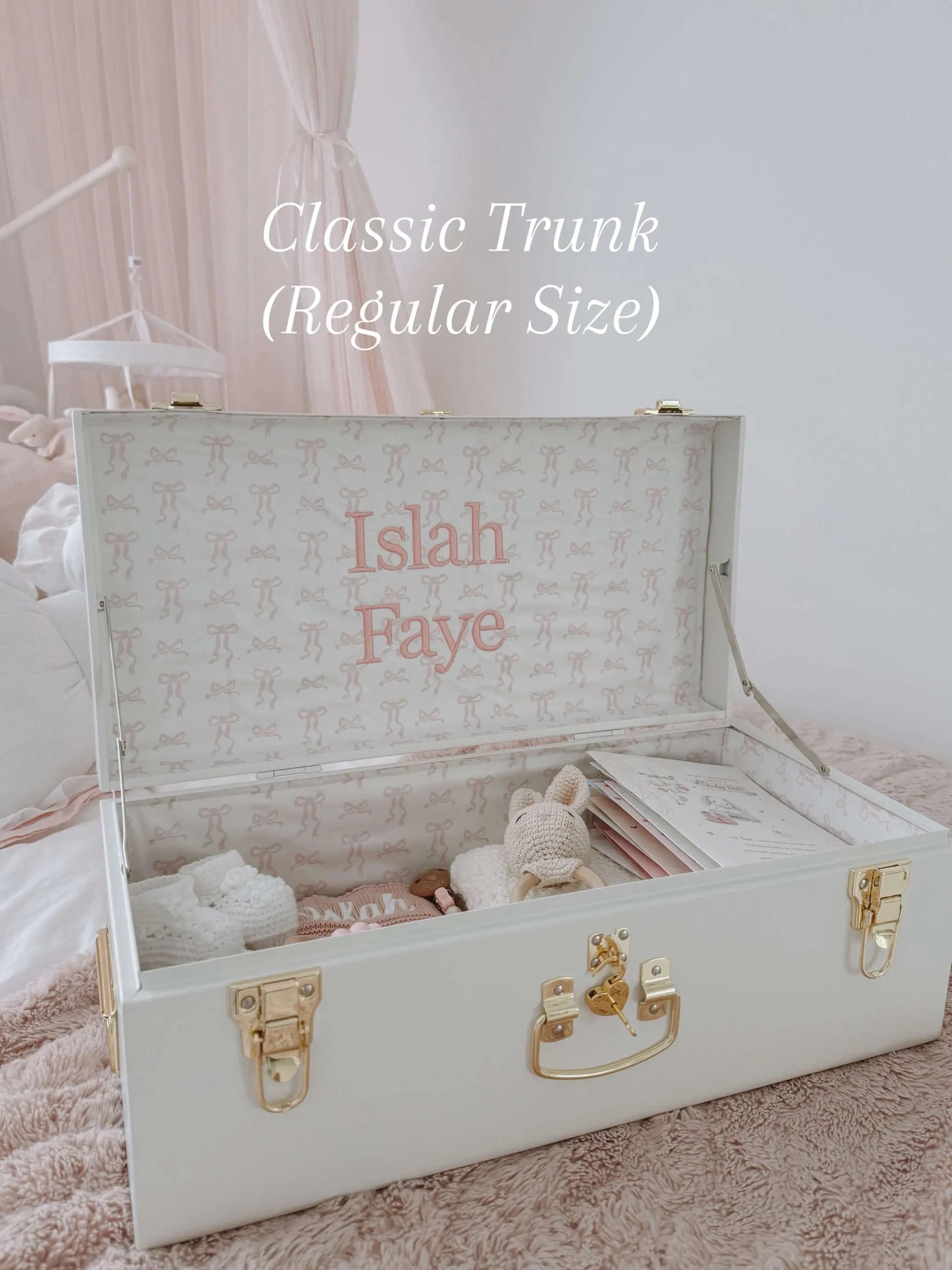 White trunk with gold accents and embroidered names, set against a soft pink background.