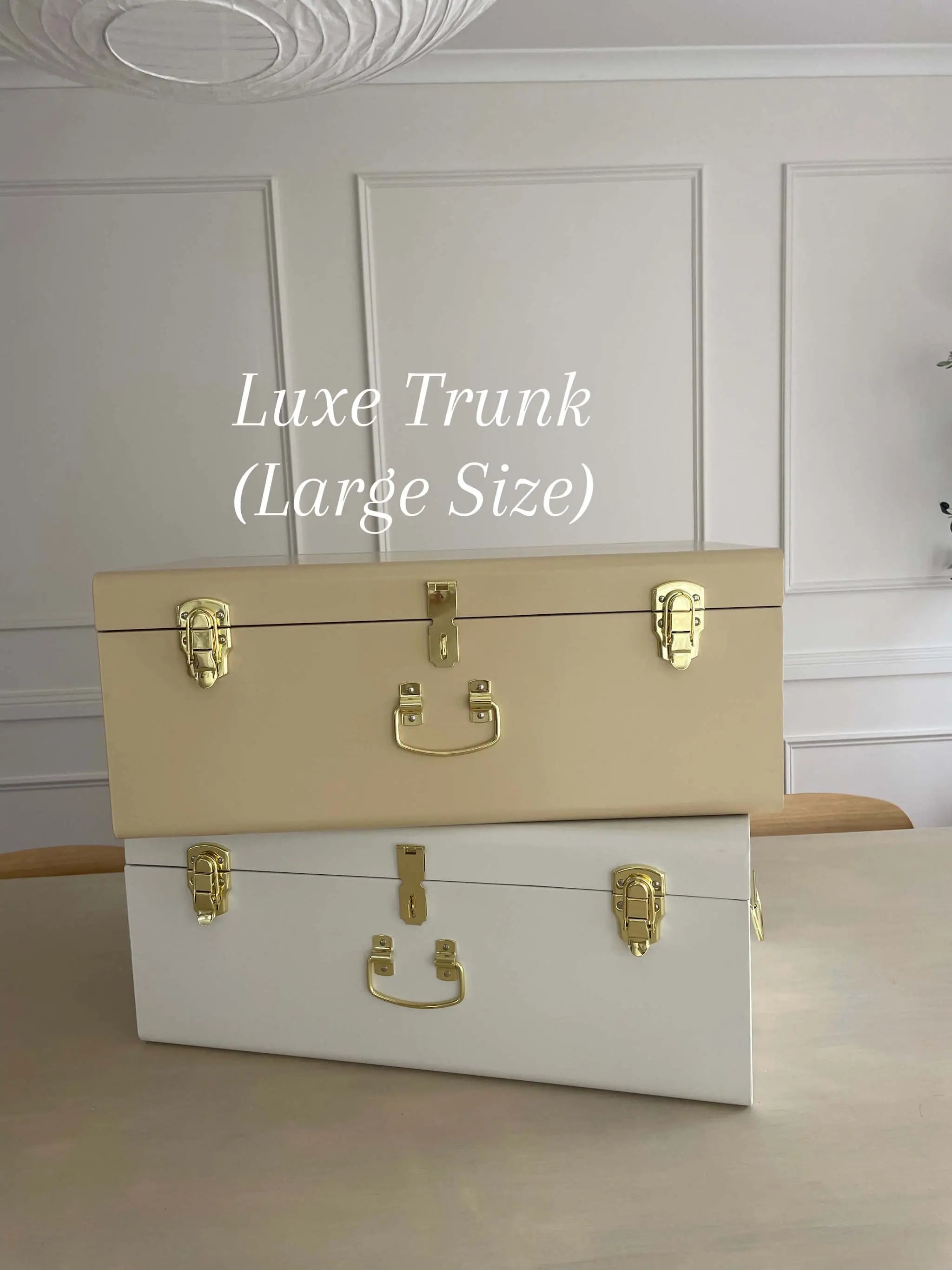 Two trunks, one beige and one white, with gold hardware on a wooden surface.