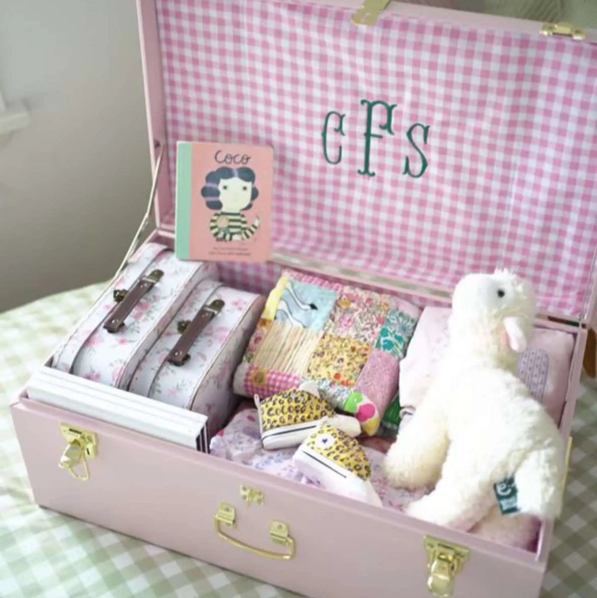 Pink gingham trunk with monogrammed initials, toys, and sewing supplies on a checkered surface.