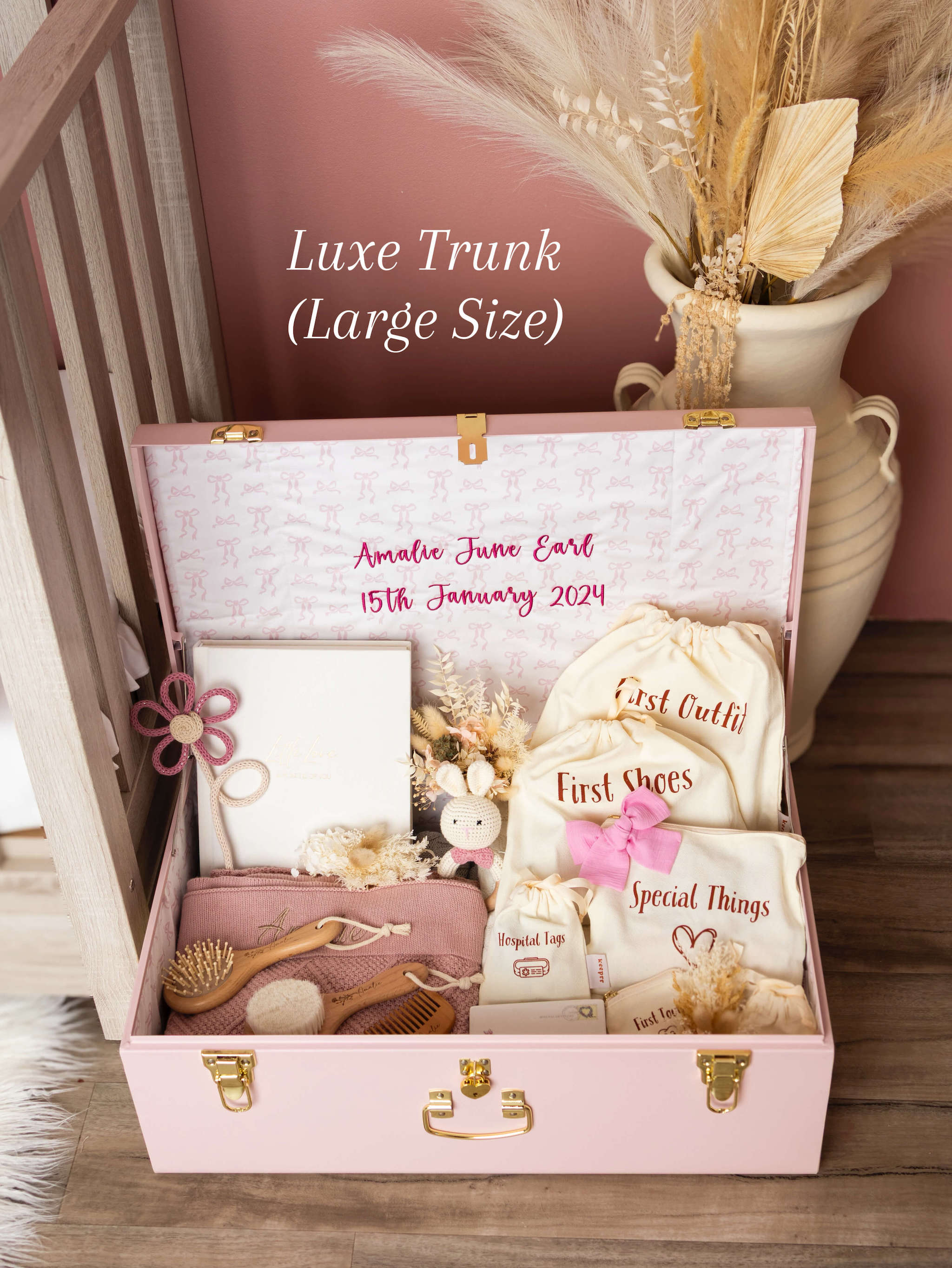 Pink baby trunk with baby items and personalized text, placed on a wooden floor.