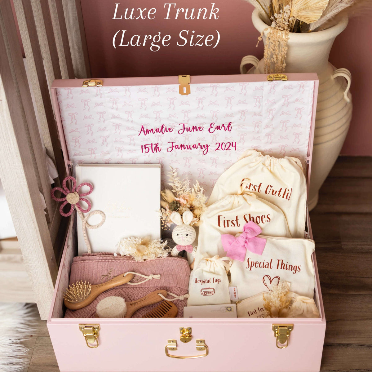 Pink baby trunk with baby items and personalized text, placed on a wooden floor.