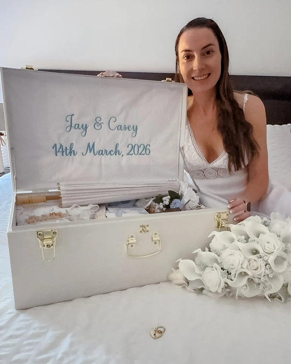 Woman holding a white wedding dress in an open suitcase with embroidered names and date, sitting on a bed.