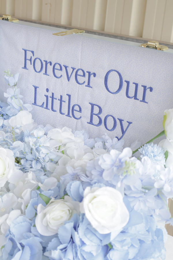 Memorial sign with 'Forever Our Little Boy' text surrounded by blue and white flowers.