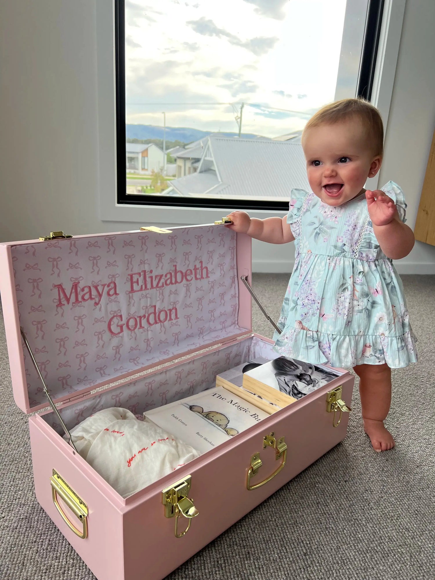 Baby in a light blue dress standing next to an open pink suitcase with &